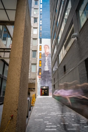 Urban view captures a tall building with a mural of a person playing violin, positioned in a narrow walkway between buildings.