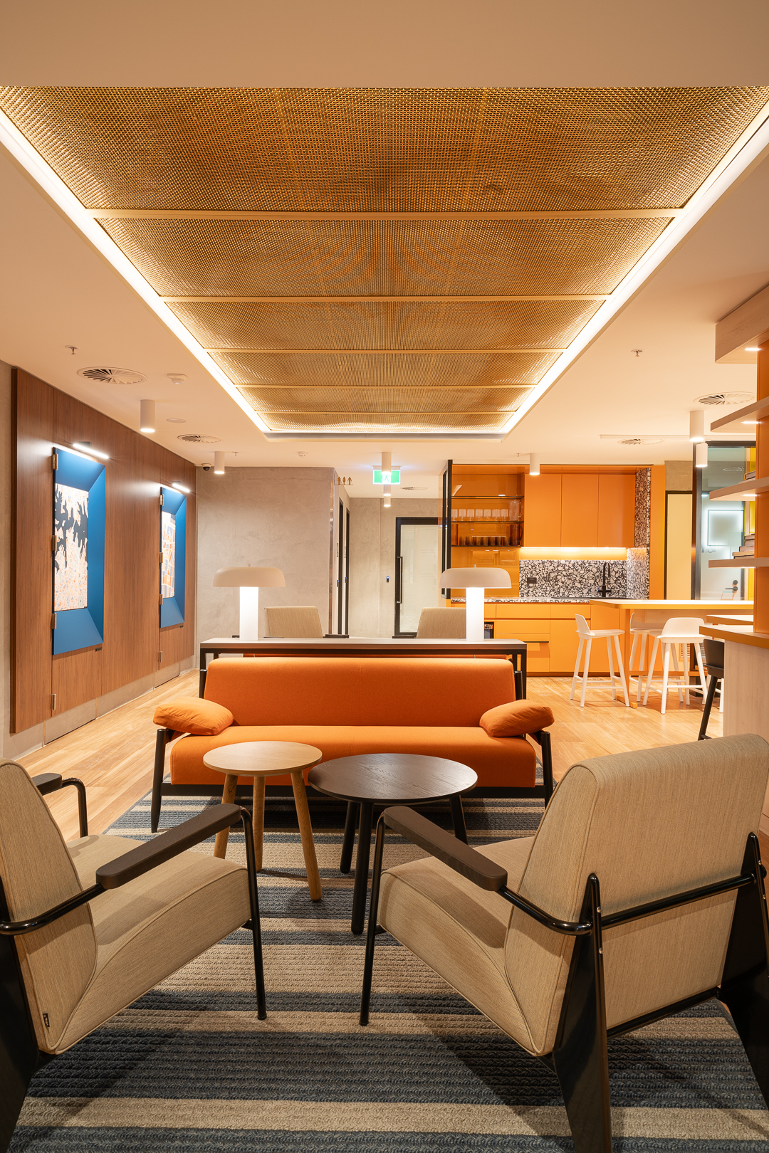 Contemporary office lounge featuring an orange couch, two armchairs, round tables, and striking ceiling lights, creating a modern, inviting space.