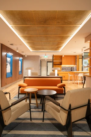 Contemporary office lounge featuring an orange couch, two armchairs, round tables, and striking ceiling lights, creating a modern, inviting space.