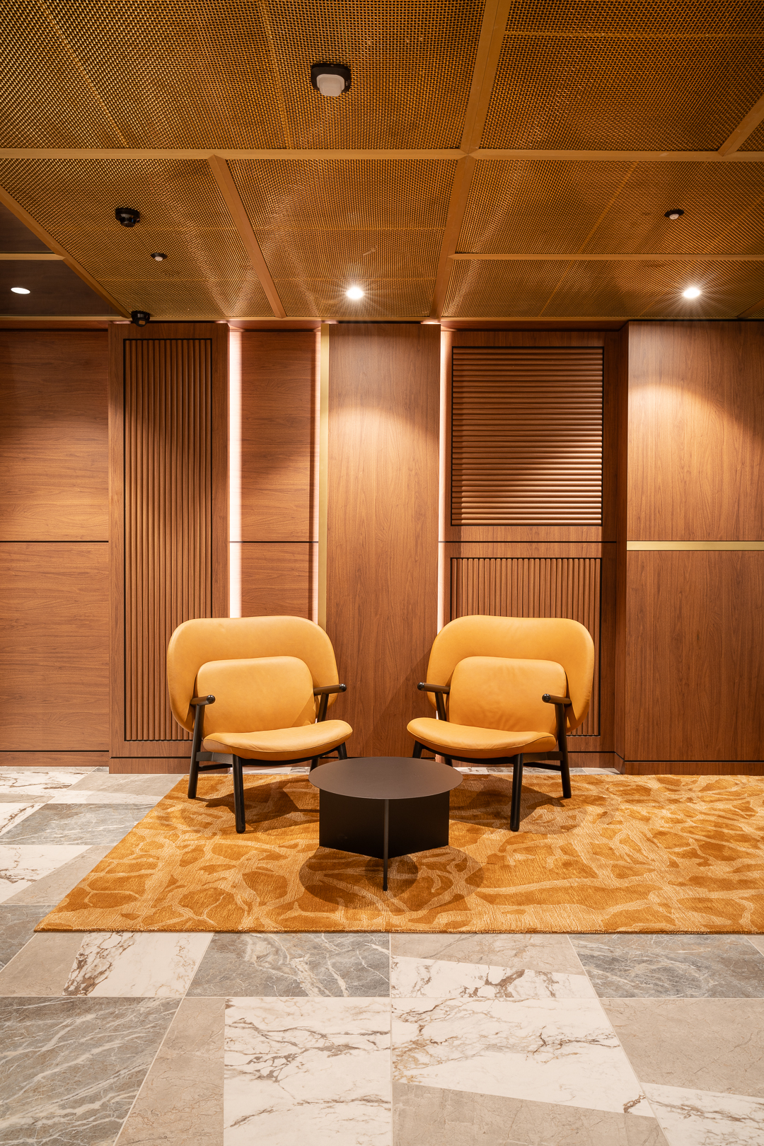 Interior view of modern lobby with two yellow armchairs, a black coffee table, marble floors and paneled wood walls, warm lighting.