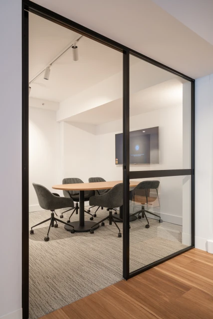Conference room with a light wood oval table and black rolling chairs seen through a black frame glass wall and door.