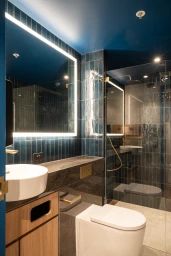 Modern commercial bathroom with blue vertical wall tiles, illuminated rectangular mirror, white vessel sink, and toilet by Intermain