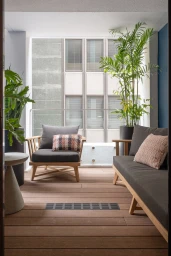 Modern outdoor balcony interior with wooden flooring, grey sofa and armchair, potted plants, and city building facade in the background
