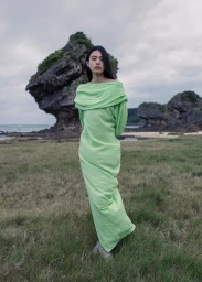 Model Kinari Tamukai in a light green off-the-shoulder dress standing on a grassy coastal landscape before rock formations.