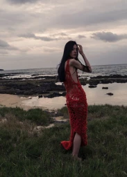 Model Kinari Tamukai wearing a red knitted dress standing on a coastal rocky shore under an overcast sky for Sasurai