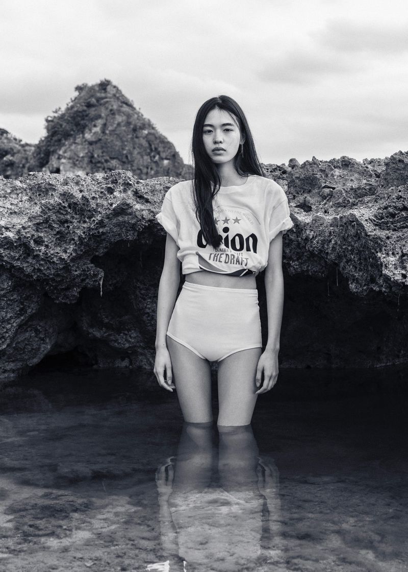 Model standing in shallow water against rocky coastal background for Sasurai editorial by 5ELEVEN Magazine