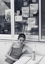 Model seated at an outdoor table wearing a patterned swimsuit and cap in front of a shop window with Japanese signage.