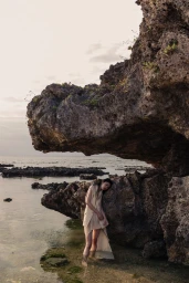 Model standing in shallow coastal water leaning against a large rocky outcropping under an overcast sky.