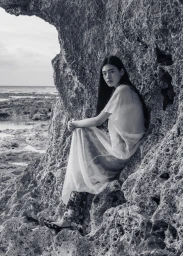 Model Kinari Tamukai seated within a rocky cave formation overlooking the ocean, wearing a sheer layered dress.