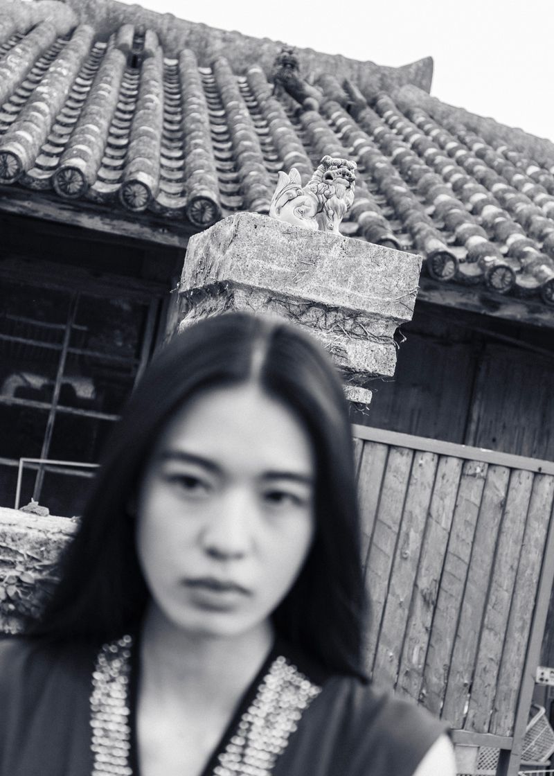 Portrait of a woman with long hair in front of a building with a tiled roof and a lion statue, captured in black and white.