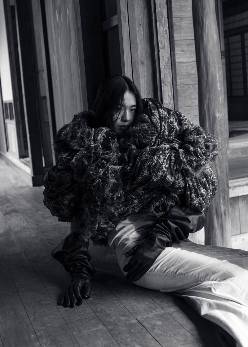 A solitary Asian woman models an oversized, textured coat with latex gloves in a hallway with a muted black and white color palette.