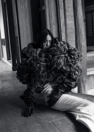 A solitary Asian woman models an oversized, textured coat with latex gloves in a hallway with a muted black and white color palette.