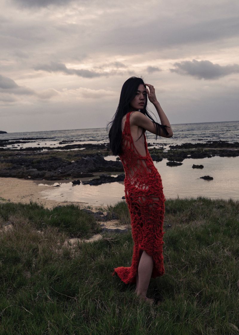 A slender woman in a red crocheted dress poses on a grassy bank overlooking the ocean with a cloudy sky at dusk or dawn.
