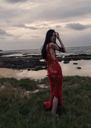 A slender woman in a red crocheted dress poses on a grassy bank overlooking the ocean with a cloudy sky at dusk or dawn.