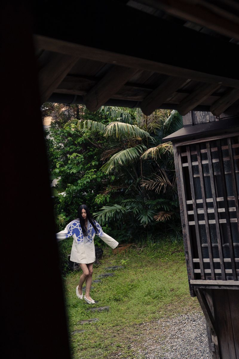 Woman in blue and white patterned top and light skirt walks on a stone path through a tropical-looking garden, seen from a dark window.