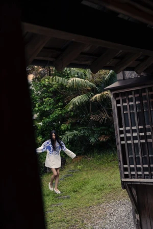 Woman in blue and white patterned top and light skirt walks on a stone path through a tropical-looking garden, seen from a dark window.