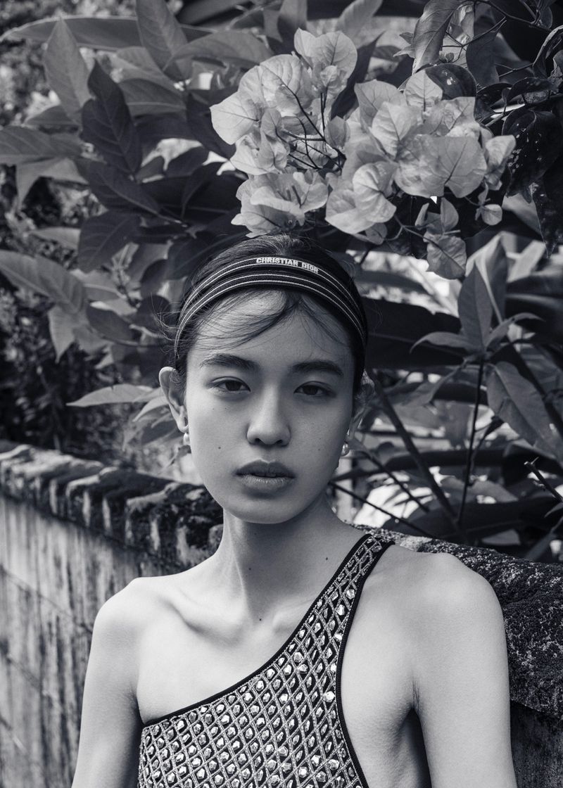 Portrait of an Asian woman wearing a patterned headband and shoulder-baring dress, posing in front of bougainvillea and a weathered wall.