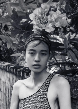 Portrait of an Asian woman wearing a patterned headband and shoulder-baring dress, posing in front of bougainvillea and a weathered wall.