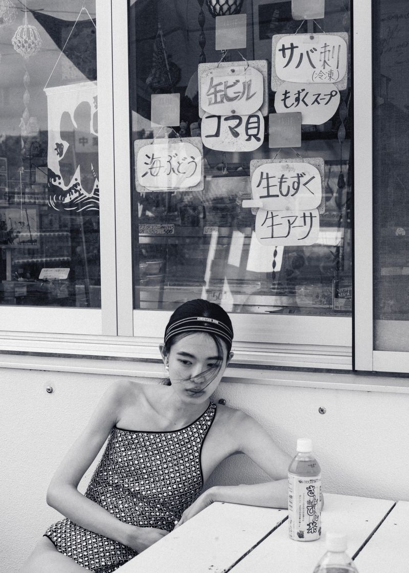 Young woman in one-shoulder swimsuit sits at white table outside, a window behind her with signs and sunlight casting shadows.