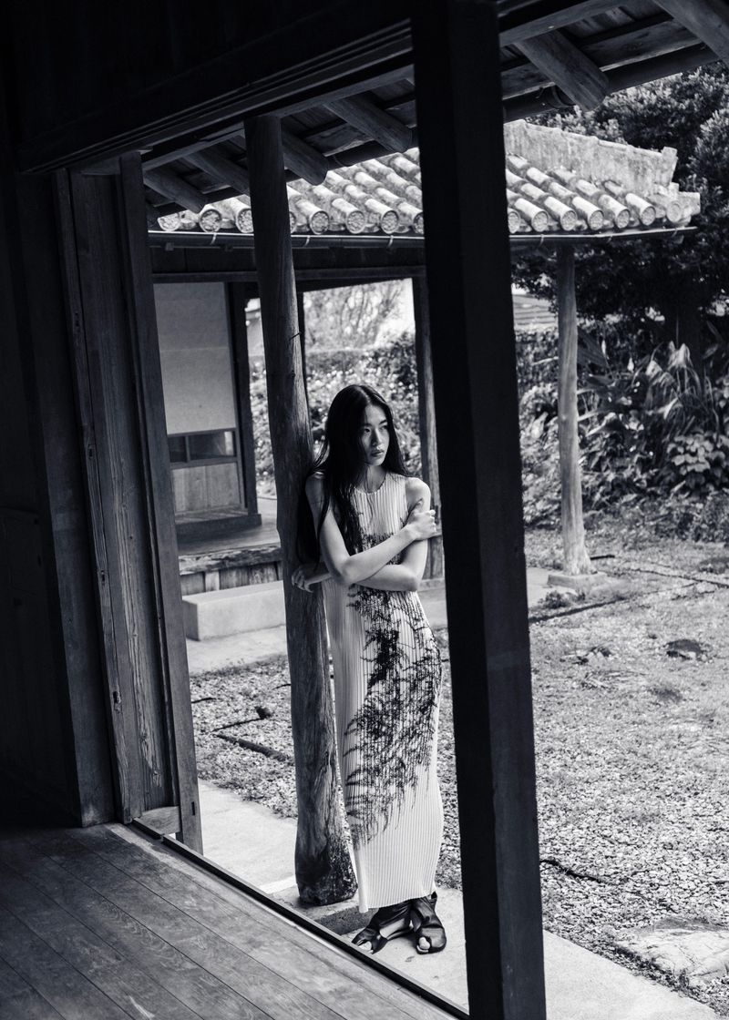 Monochrome full shot of a woman in a patterned dress standing in a traditional Japanese doorway looking toward the garden.