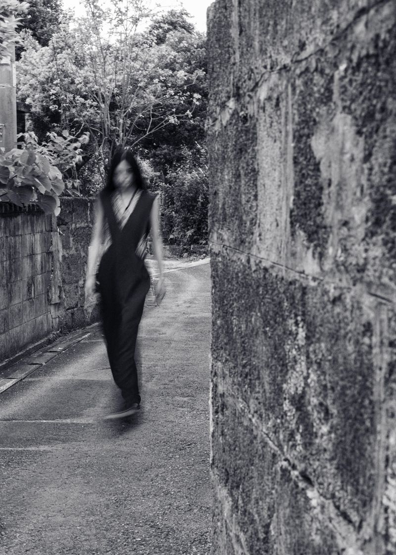 Monochrome shot of an adult female walking on a path lined with brick walls and trees in the background, creating a blurred motion effect.