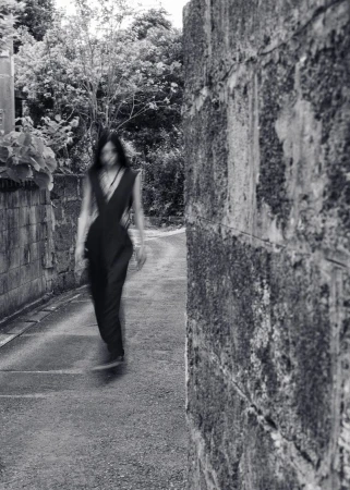 Monochrome shot of an adult female walking on a path lined with brick walls and trees in the background, creating a blurred motion effect.