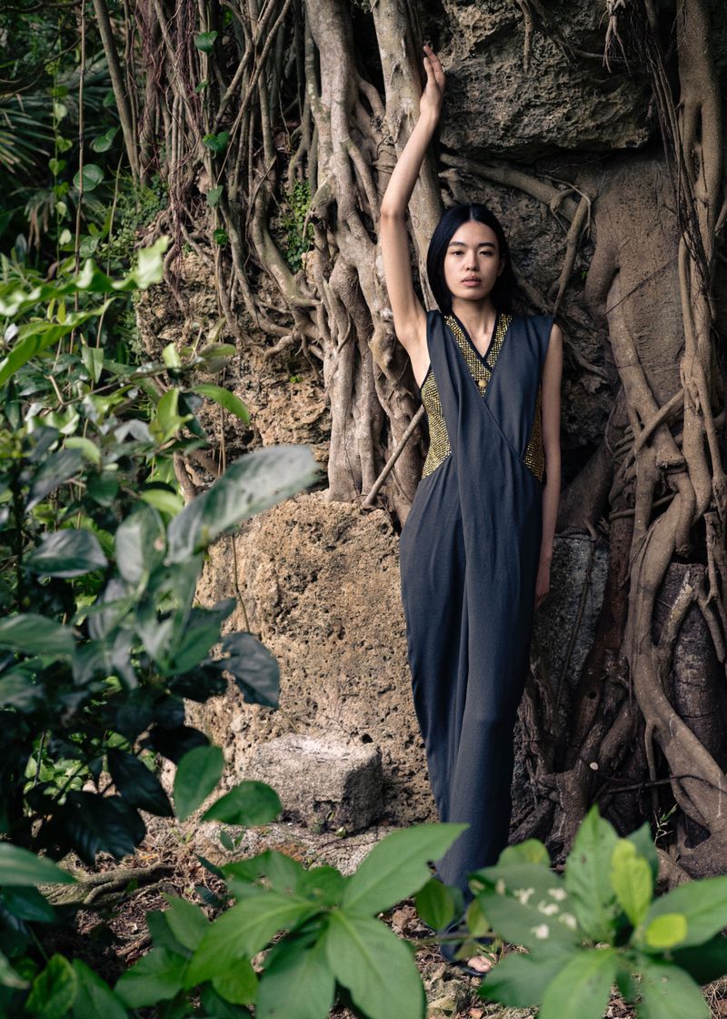 Adult woman in an elegant grey dress with gold accents poses next to a tree with exposed roots in a lush tropical forest setting.
