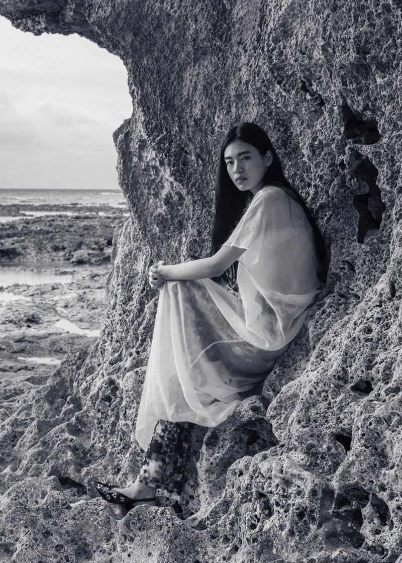 Asian woman sits on rocky beach outcrop near ocean in sheer dress, floral leggings, and black shoes, gazing at the camera in monochrome.