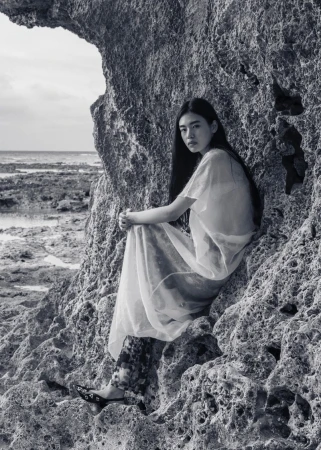 Asian woman sits on rocky beach outcrop near ocean in sheer dress, floral leggings, and black shoes, gazing at the camera in monochrome.