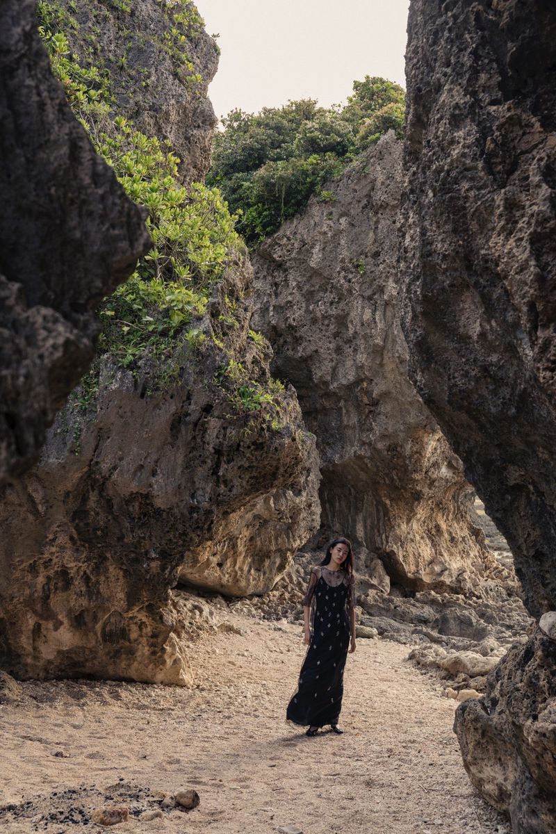 Adult female model in long dress standing on sand path between eroded rock formations in a natural outdoor setting with green foliage.