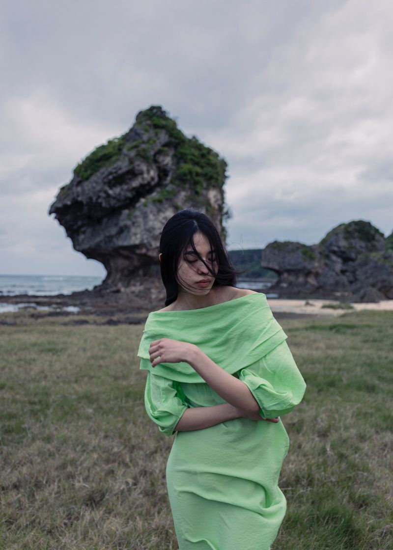 A woman in a green off-the-shoulder dress stands on a grassy beach with an interesting rock formation and ocean in the background.