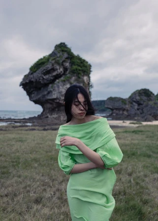 A woman in a green off-the-shoulder dress stands on a grassy beach with an interesting rock formation and ocean in the background.