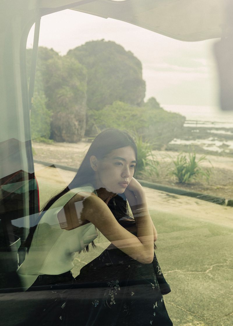 Pensive young woman gazes from a car window at a coastal landscape featuring a beach, ocean, and lush foliage-covered rocky cliffs.