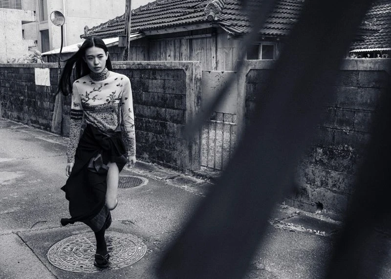 Monochrome fashion shot of an Asian woman in a patterned top and skirt, walking on a city street in front of a textured wall and buildings.