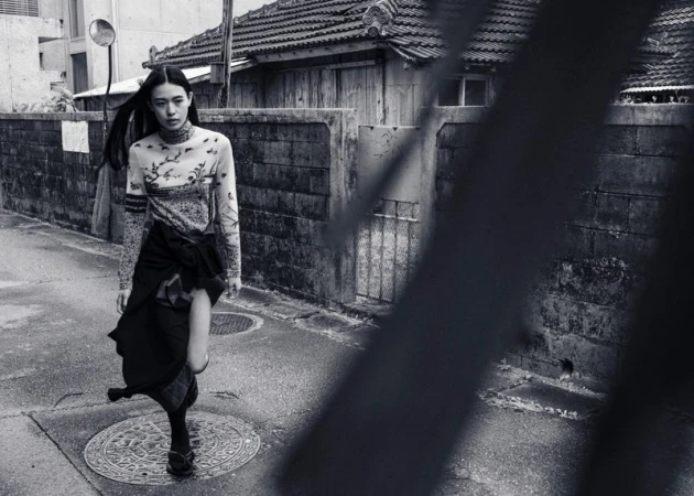 Monochrome fashion shot of an Asian woman in a patterned top and skirt, walking on a city street in front of a textured wall and buildings.