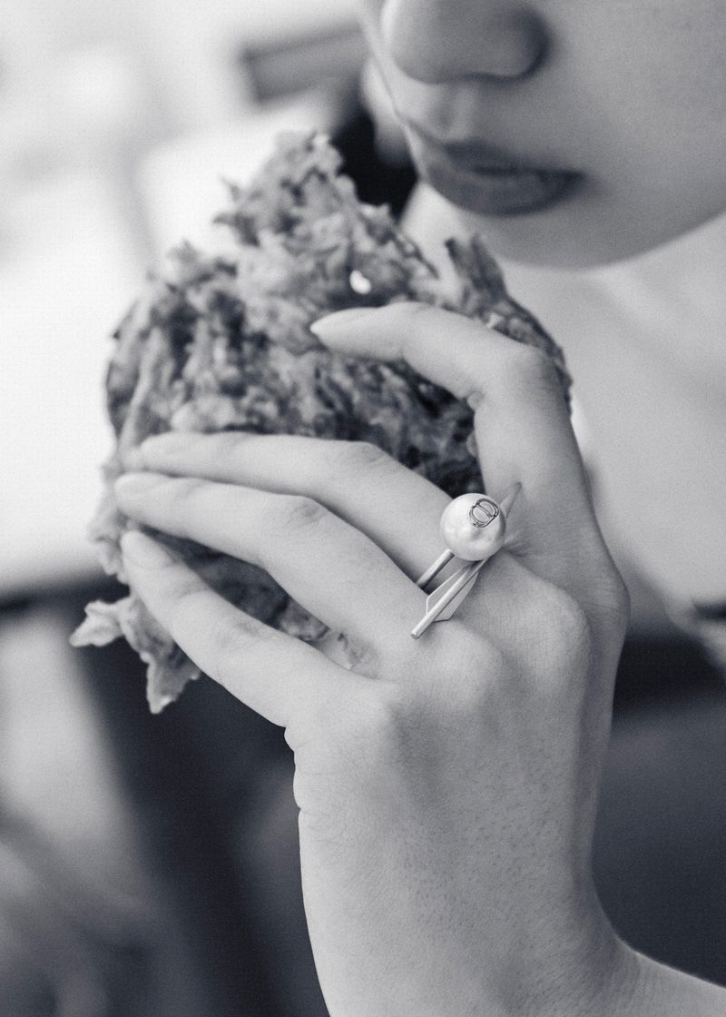 Black and white close-up of a person holding and eating a fried food item, featuring a silver ring with a pearl accent.