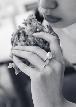 Black and white close-up of a person holding and eating a fried food item, featuring a silver ring with a pearl accent.