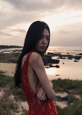 Portrait of an Asian woman wearing a red macrame dress, standing on a grassy shore with the ocean and a cloudy sky in the background.