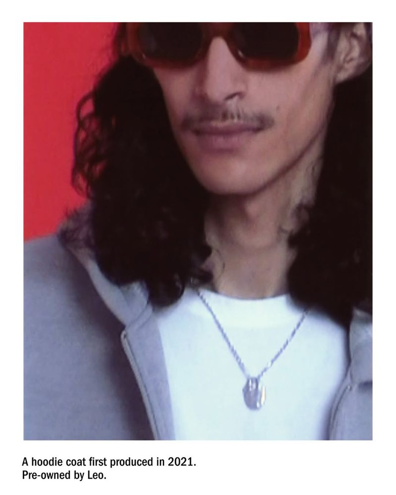 Close-up studio shot of a man with long hair wearing sunglasses, a necklace, and a hoodie against a red backdrop.