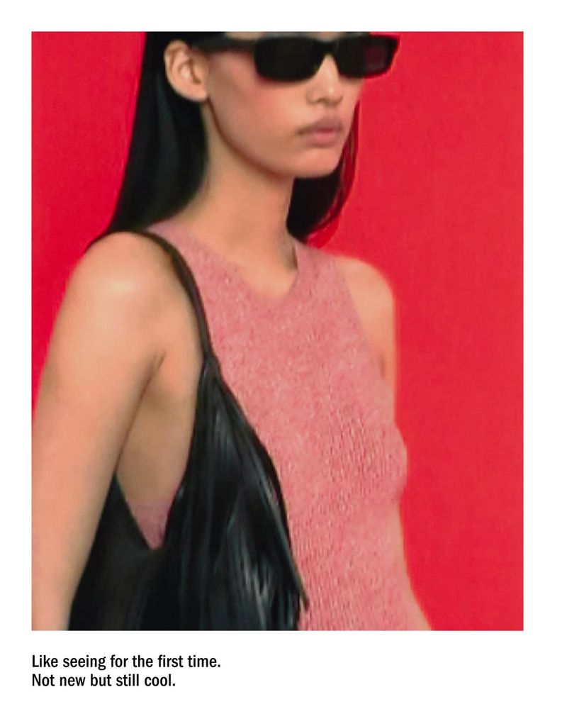 Model wearing pink knit top and black sunglasses holding a black handbag against a solid red backdrop
