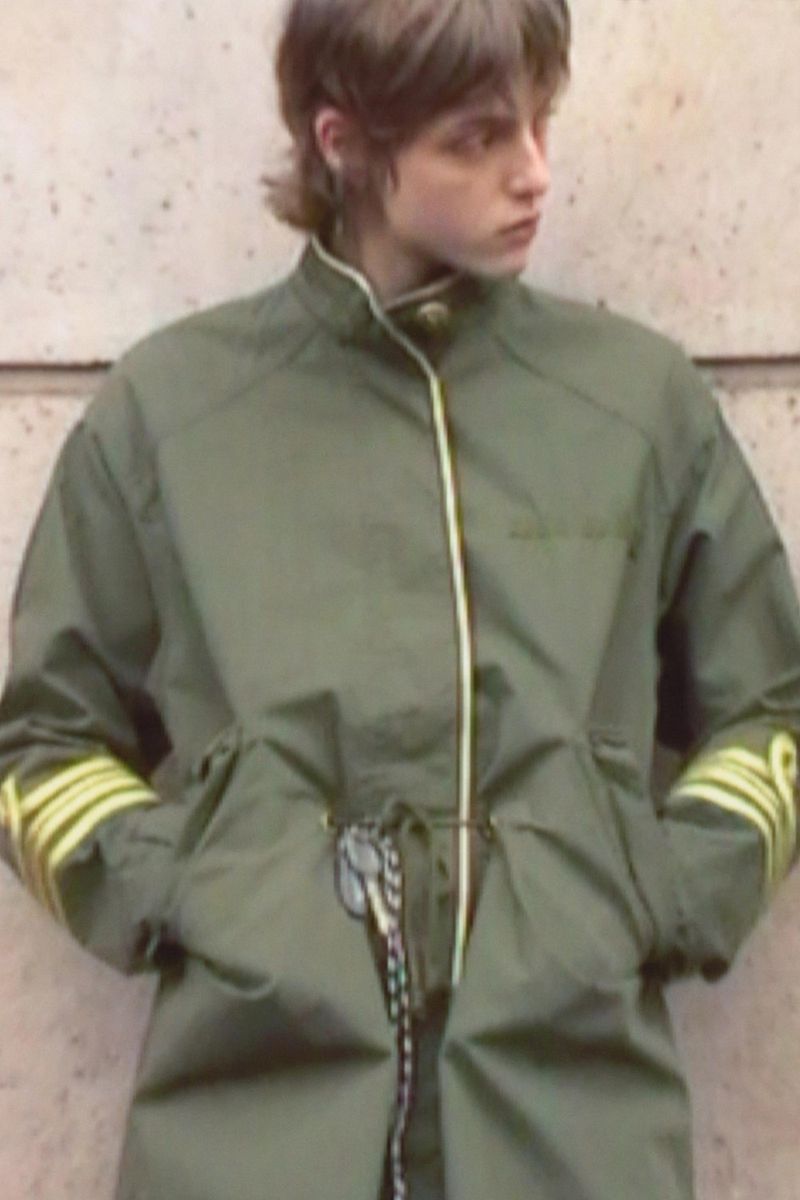 Model wearing an olive green jacket with yellow striped sleeve details standing against a stone wall