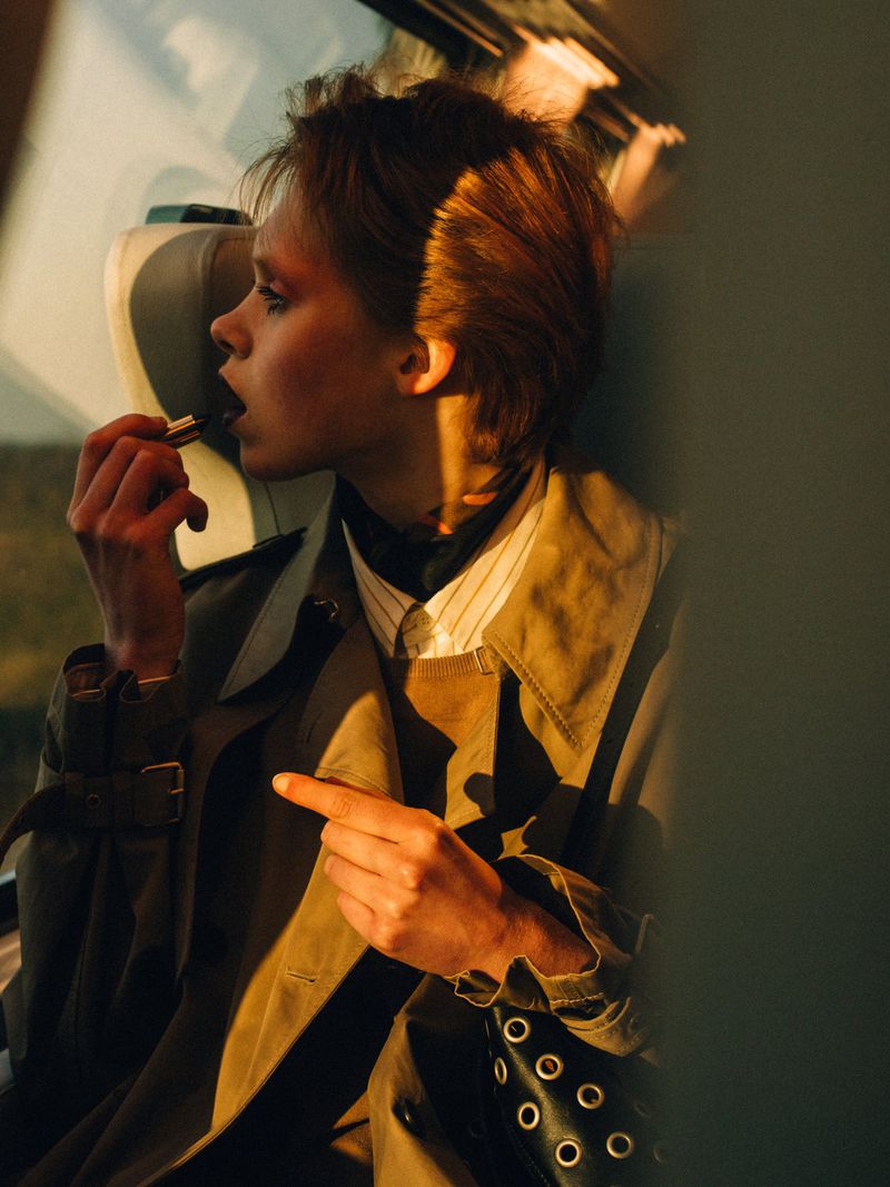 Young woman applies lipstick inside a car in warm light, wearing a trench coat, highlighting a beauty shot emphasizing makeup application.