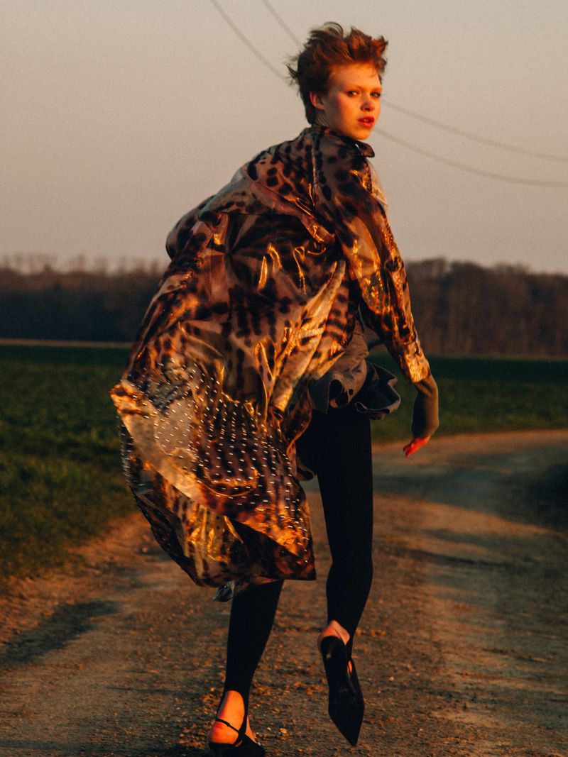Outdoor fashion shot: a person in a patterned coat walks on a dirt path with a field and trees in the background at sunset.