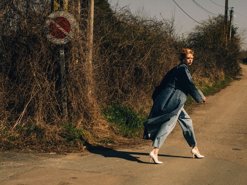 Fashionable young adult female strides confidently on rural road in a coat and heels, by a no-entry sign overgrown with branches.