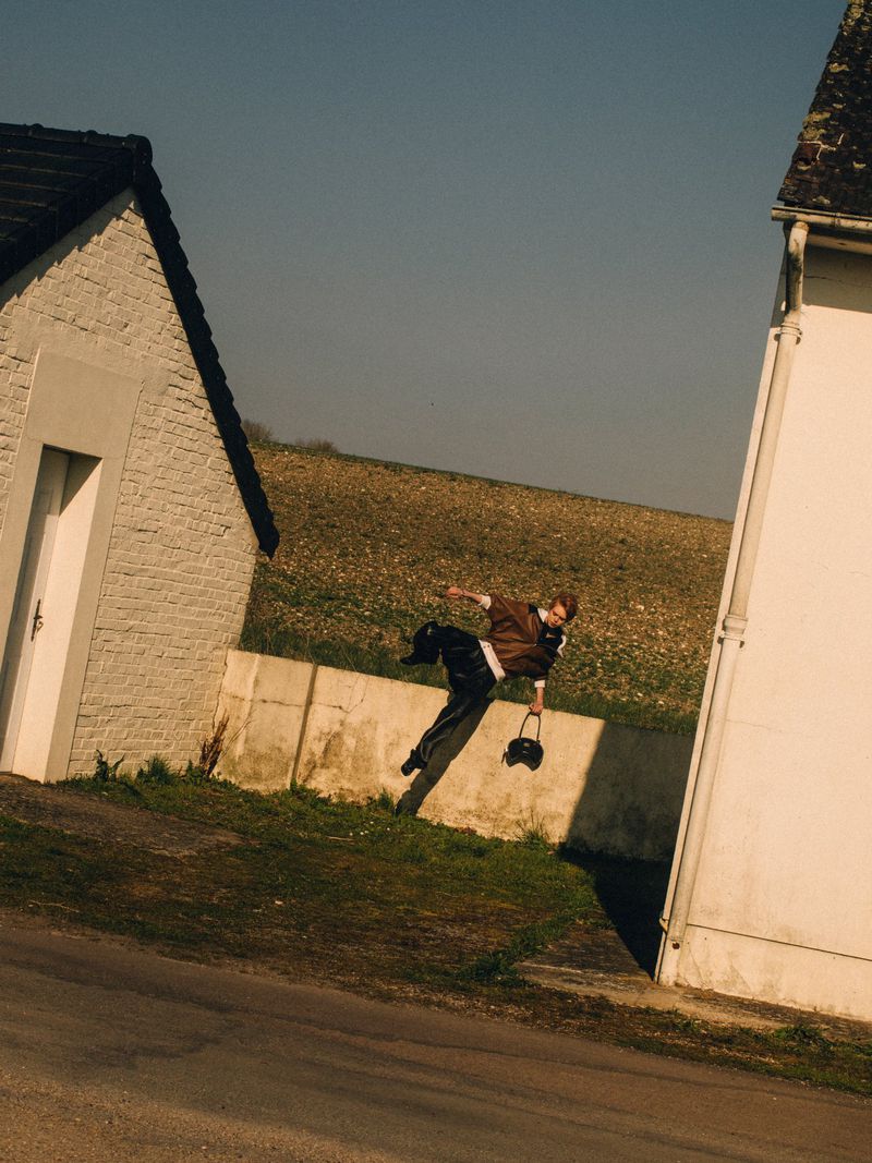 A man is suspended in mid-air between two buildings in a rural area, captured at an oblique angle on a clear day, with a brown field in the background.