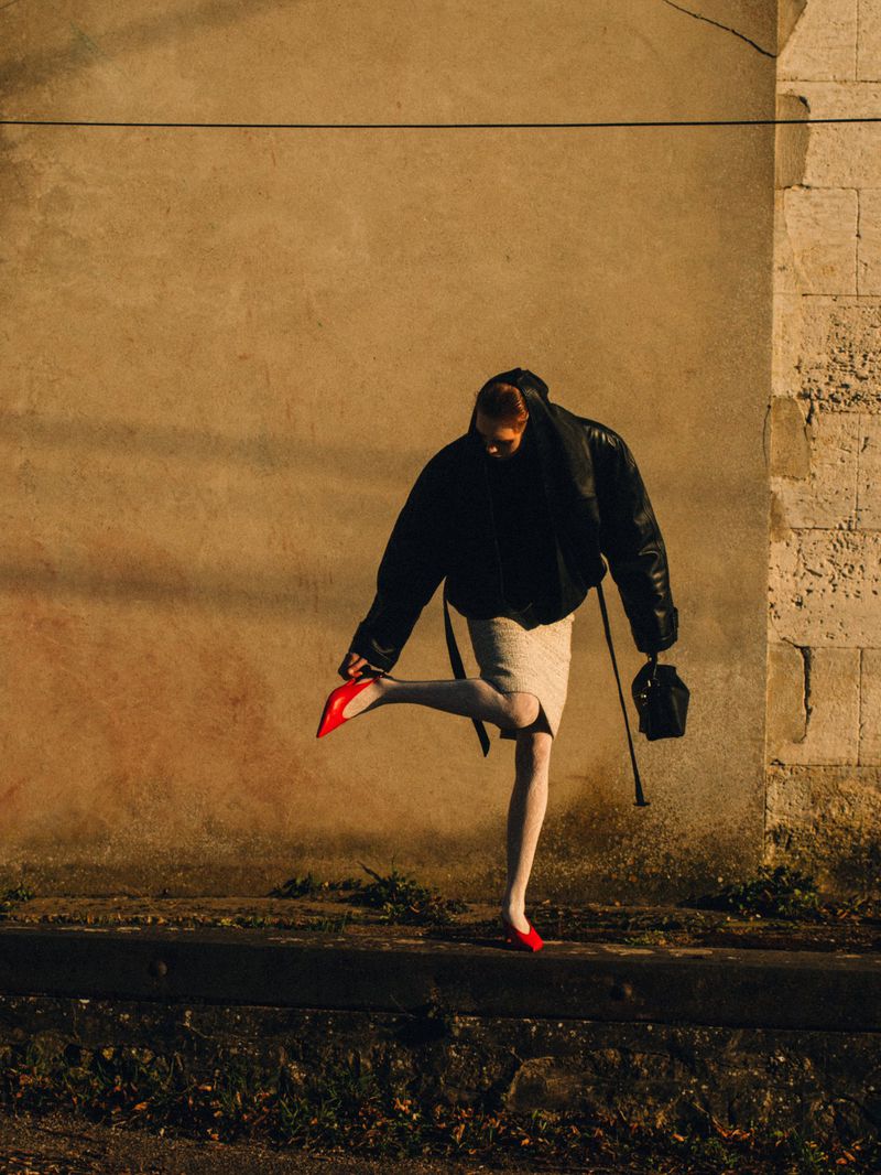 Woman in black jacket and red heels posing against a textured wall in an outdoor fashion shot. The image captures urban style.