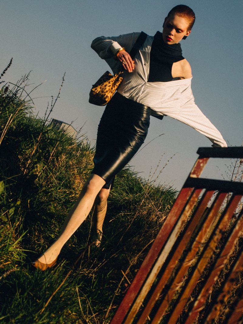 Woman poses in outdoor fashion shot wearing black skirt, striped shirt and carrying a leopard-print purse in a field of grass.