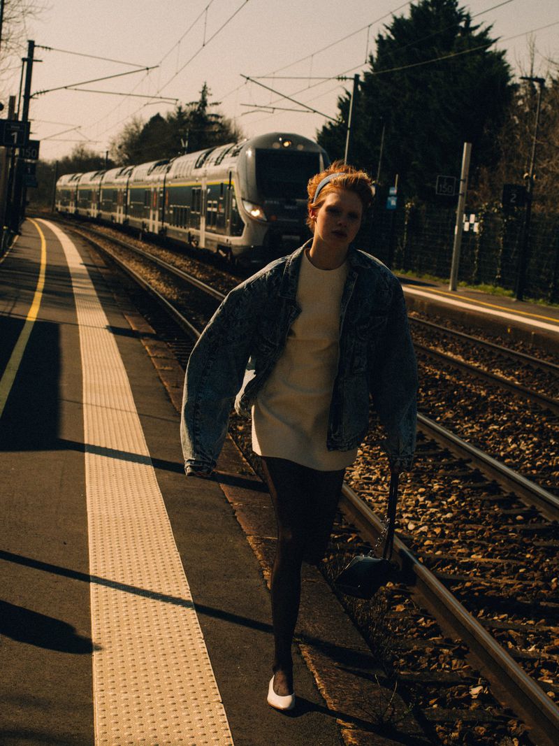 Young woman in denim jacket walks along train tracks next to a modern passenger train at an outdoor station in daylight.
