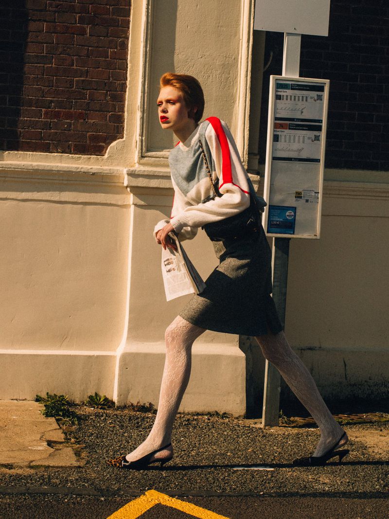 A red-haired woman with fair skin in a grey sweater and skirt walks past a bus stop wearing sheer white tights on a sunny day.