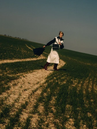 Woman poses in field wearing white apron dress, black purse, and red boots against blue sky, exuding fashion editorial style.